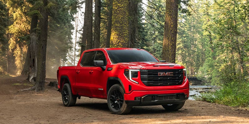 A red 2026 GMC Sierra 1500 truck is parked beside a wide creek in a forested park.