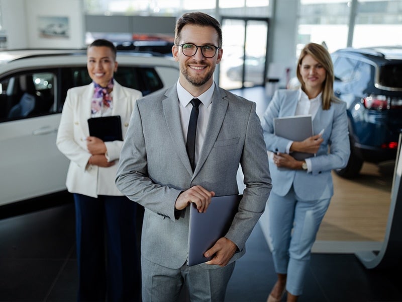 2 male and 1female sales representatives are standing among the vehicles on a sunlit GMC showroom floor, holding their Ipads and smiling at us, ready to help customers in their searches for a new vehicle.