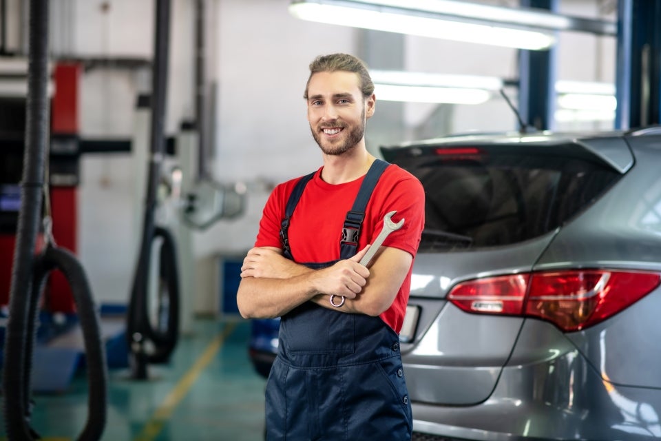 A very friendly-looking Buick and GMC Service Center mechanic in a red t-shirt and blue overalls stands among the vehicles he's inspecting and is smiling at us with his arms folded as he holds a wrench in his hand. 